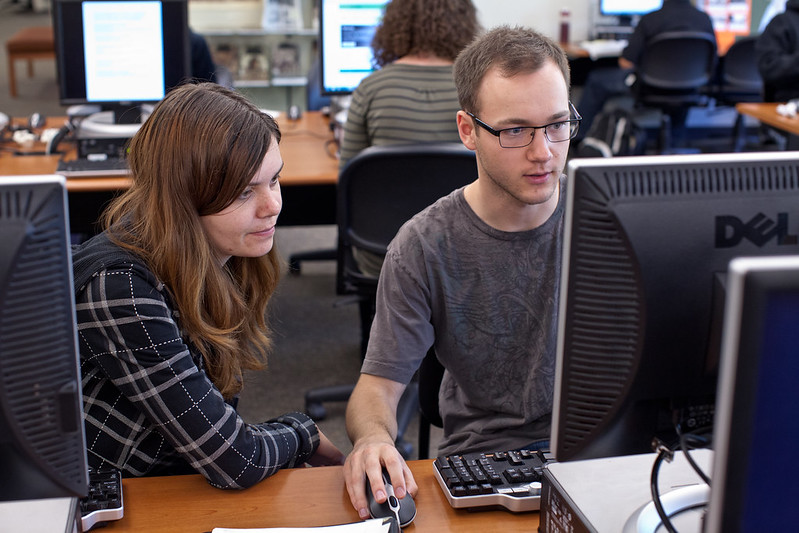 Students collaborating in a computer lab, one leans over to check another's screen.